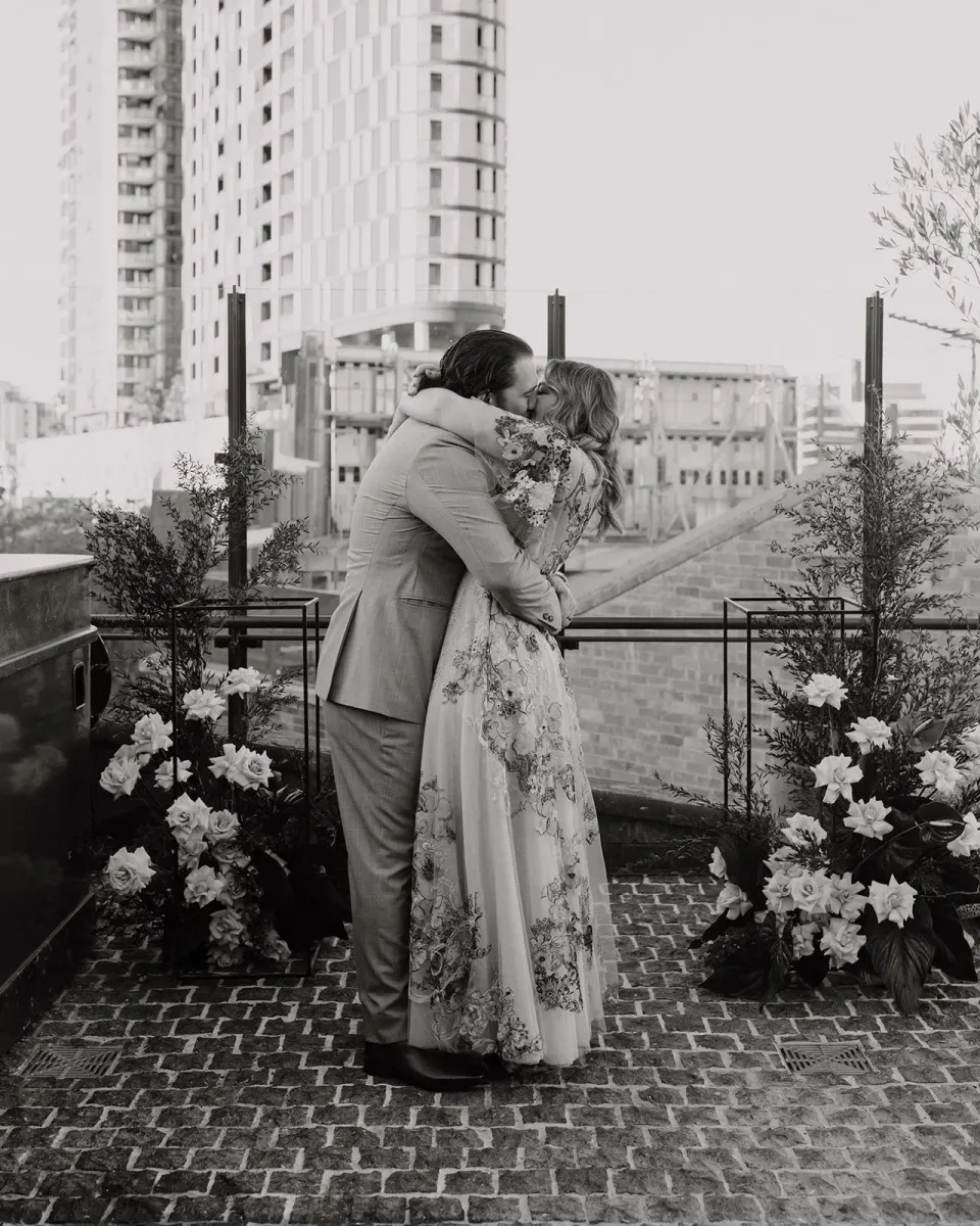 Black-and-white photograph of Victoria and Daniel kissing between ceremony flowers by Mrs Gibbons Flowers, at Agnes Restaurant in Fortitude Valley, Brisbane, for an Elopement Collective elopement. Photo by Tessa Shannon.