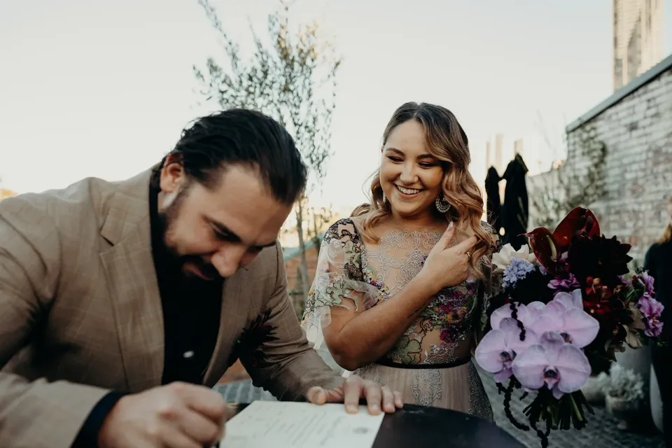 Daniel signing the marriage paperwork as Victoria laughs beside him with her bouquet by Mrs Gibbons Flowers, at Agnes Restaurant in Fortitude Valley, Brisbane, for an Elopement Collective elopement. Photo by Tessa Shannon.