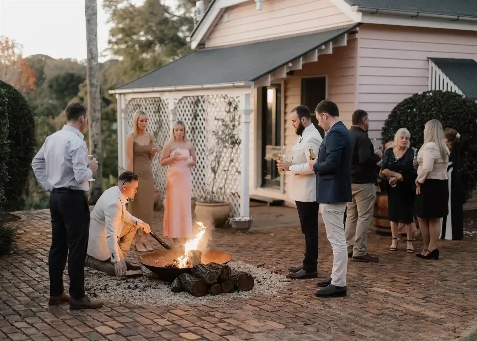 Guests gathering beside a courtyard fire outside a pale weatherboard house, in Montville, Sunshine Coast Hinterland, Queensland, for an Elopement Collective elopement. Photo by Bulb Creative.