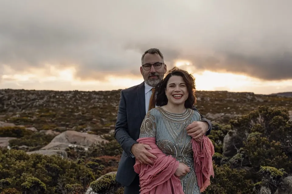 A smiling couple embrace among mountain boulders, with a pink shawl around the bride's arms.