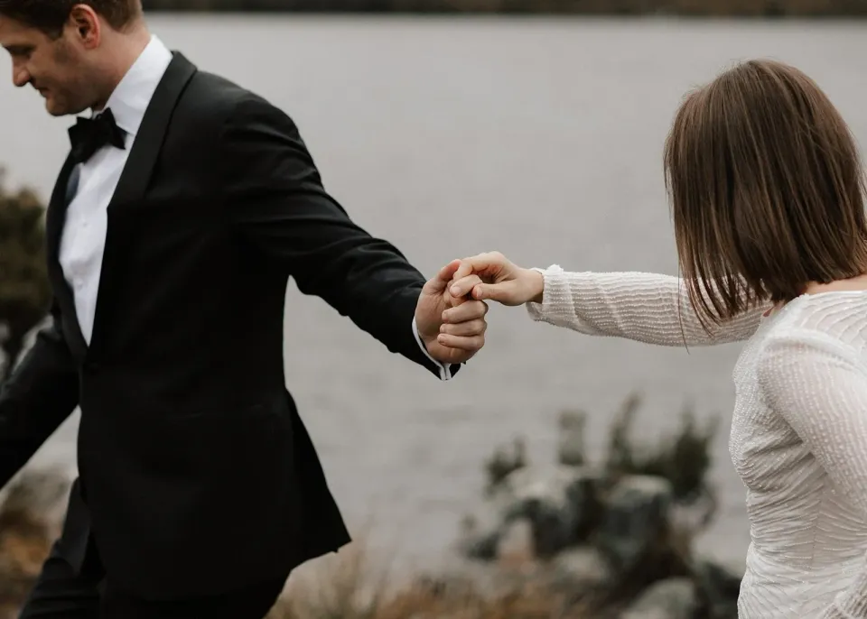 Close view of April and Josh holding hands as they walk beside the lake, at Dove Lake, Cradle Mountain, Tasmania, for an Elopement Collective elopement. Photo by Bulb Creative.