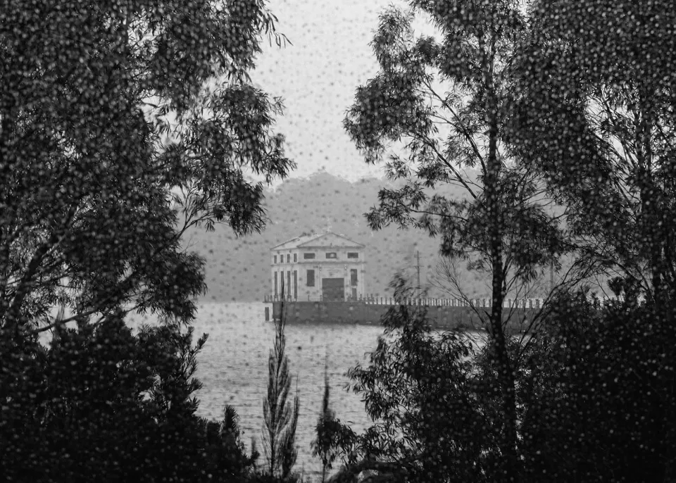Black-and-white view of the Pumphouse building across the lake, framed by leafy trees, at Pumphouse Point, Lake St Clair, Tasmania, for an Elopement Collective elopement. Photo by Bulb Creative.