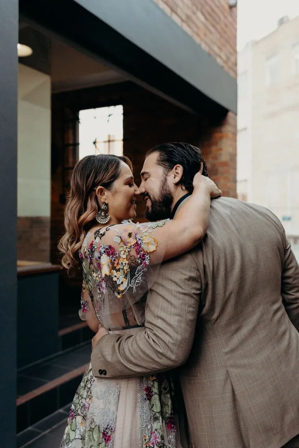 Victoria and Daniel embracing and kissing beside a dark doorway in a brick-lined laneway, in Fortitude Valley, Brisbane, for an Elopement Collective elopement. Photo by Tessa Shannon.