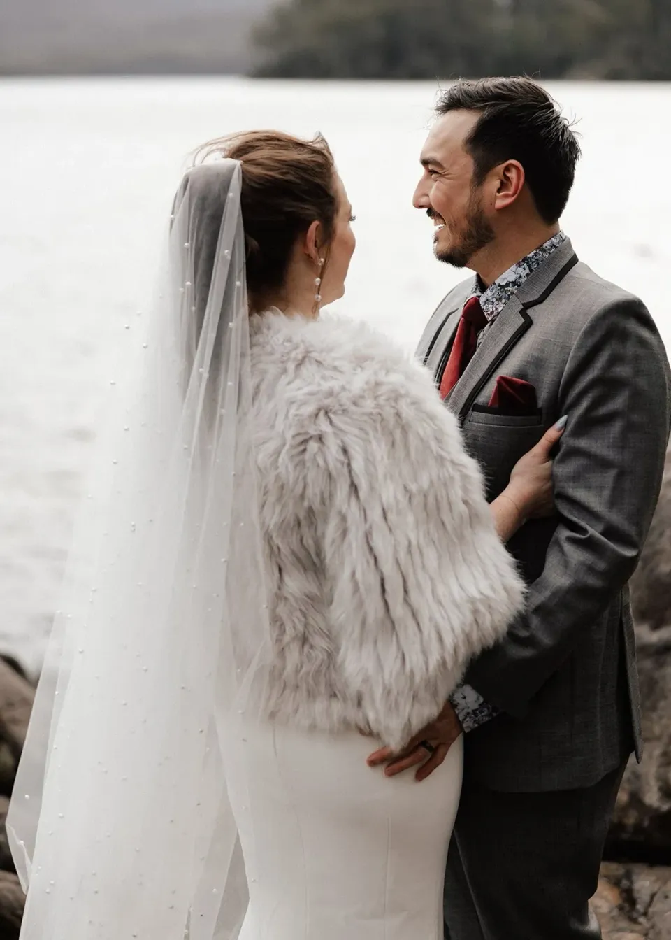 Sarah and Jono smiling together beside the lake, her wrap and long veil visible, at Pumphouse Point, Lake St Clair, Tasmania, for an Elopement Collective elopement. Photo by Bulb Creative.