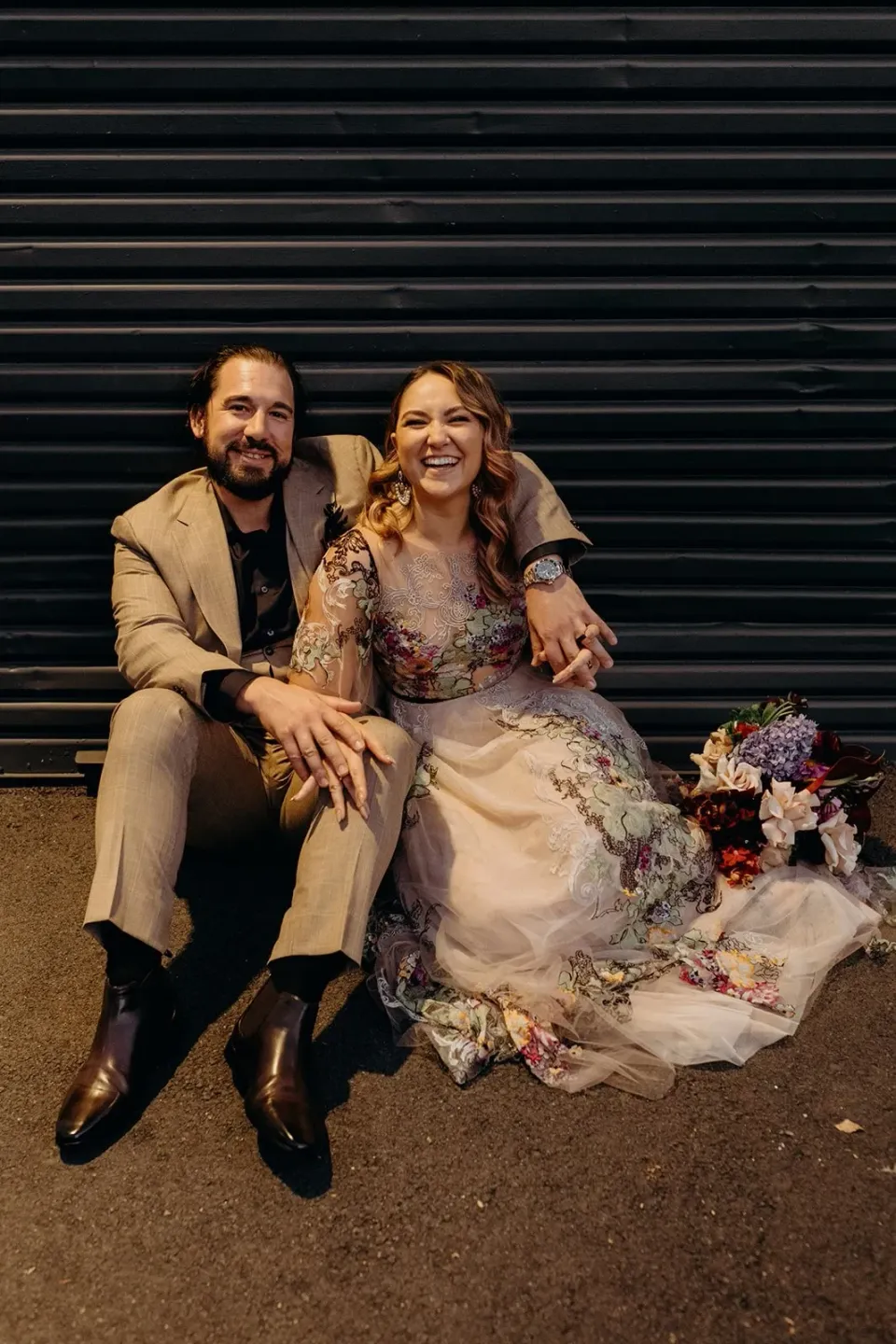 Victoria and Daniel smiling together while seated outside a black roller door, with her bouquet by Mrs Gibbons Flowers beside them, in Fortitude Valley, Brisbane, for an Elopement Collective elopement. Photo by Tessa Shannon.
