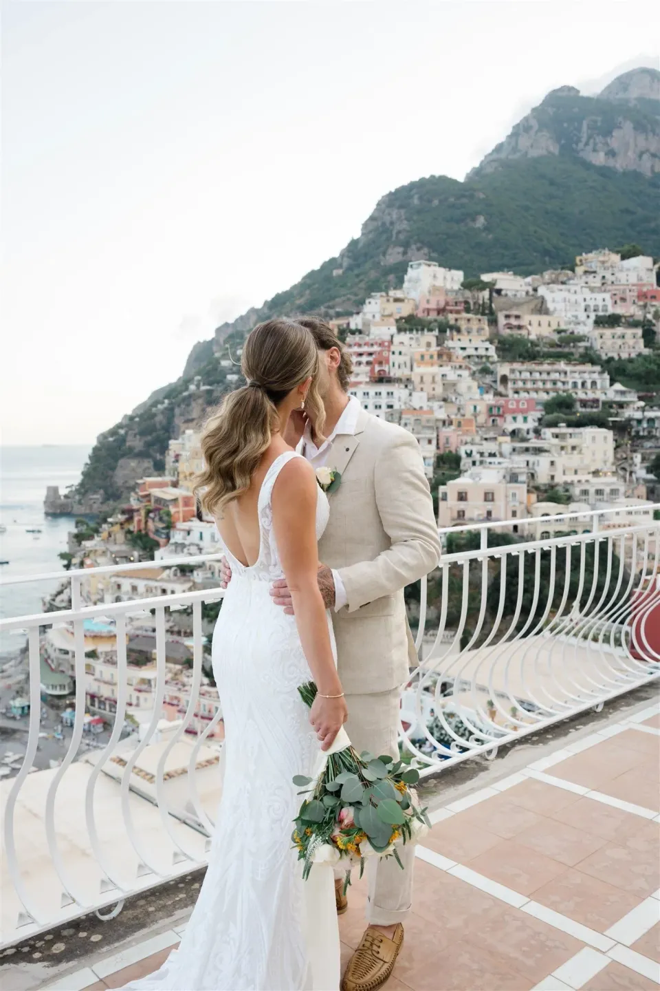 Sarah and Josh facing each other on a tiled terrace, with her bouquet by Floral Garden Positano and pastel hillside houses behind them, at Alcione Residence in Positano, Amalfi Coast, Italy, for an Elopement Collective elopement. Photo by James from House of Love.