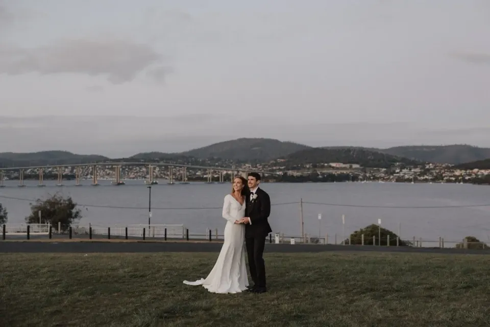 A couple stand on a lawn overlooking the river and distant hills.