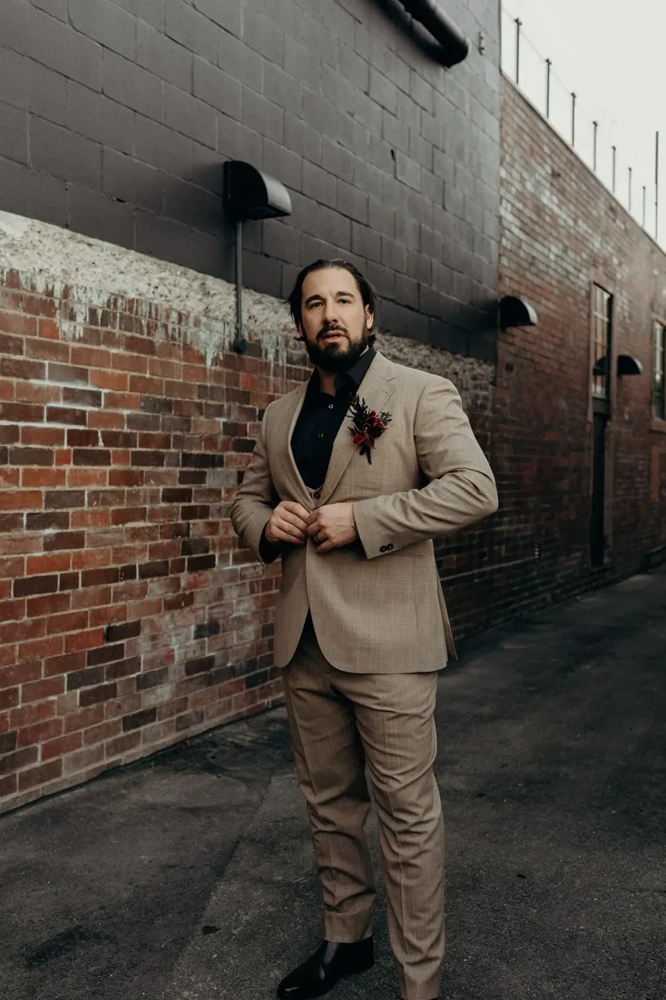 Daniel standing in a narrow brick-lined laneway, adjusting the front of his beige suit, in Fortitude Valley, Brisbane, for an Elopement Collective elopement. Photo by Tessa Shannon.