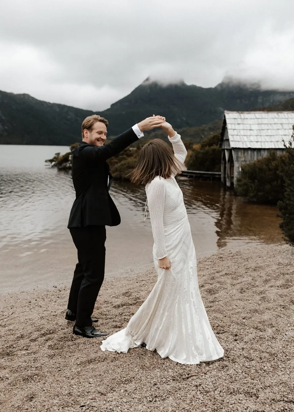 Josh twirling April on the sand beside the stone boatshed, at Dove Lake, Cradle Mountain, Tasmania, for an Elopement Collective elopement. Photo by Bulb Creative.