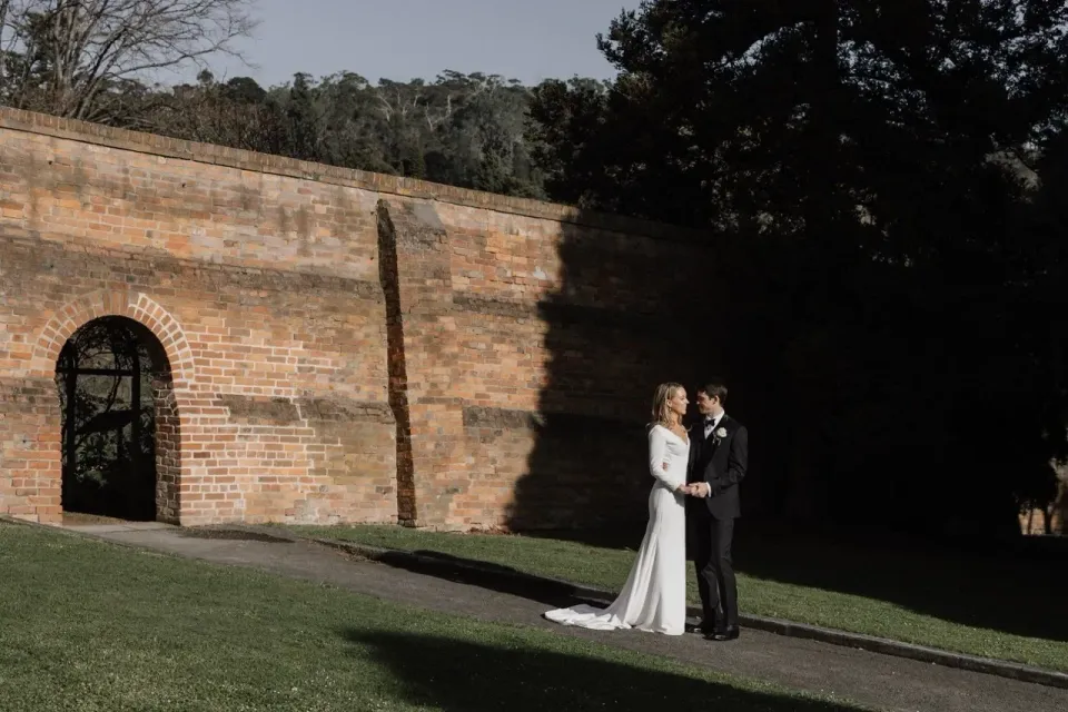 A couple stand in sunlight beside a high brick garden wall and arched gateway.