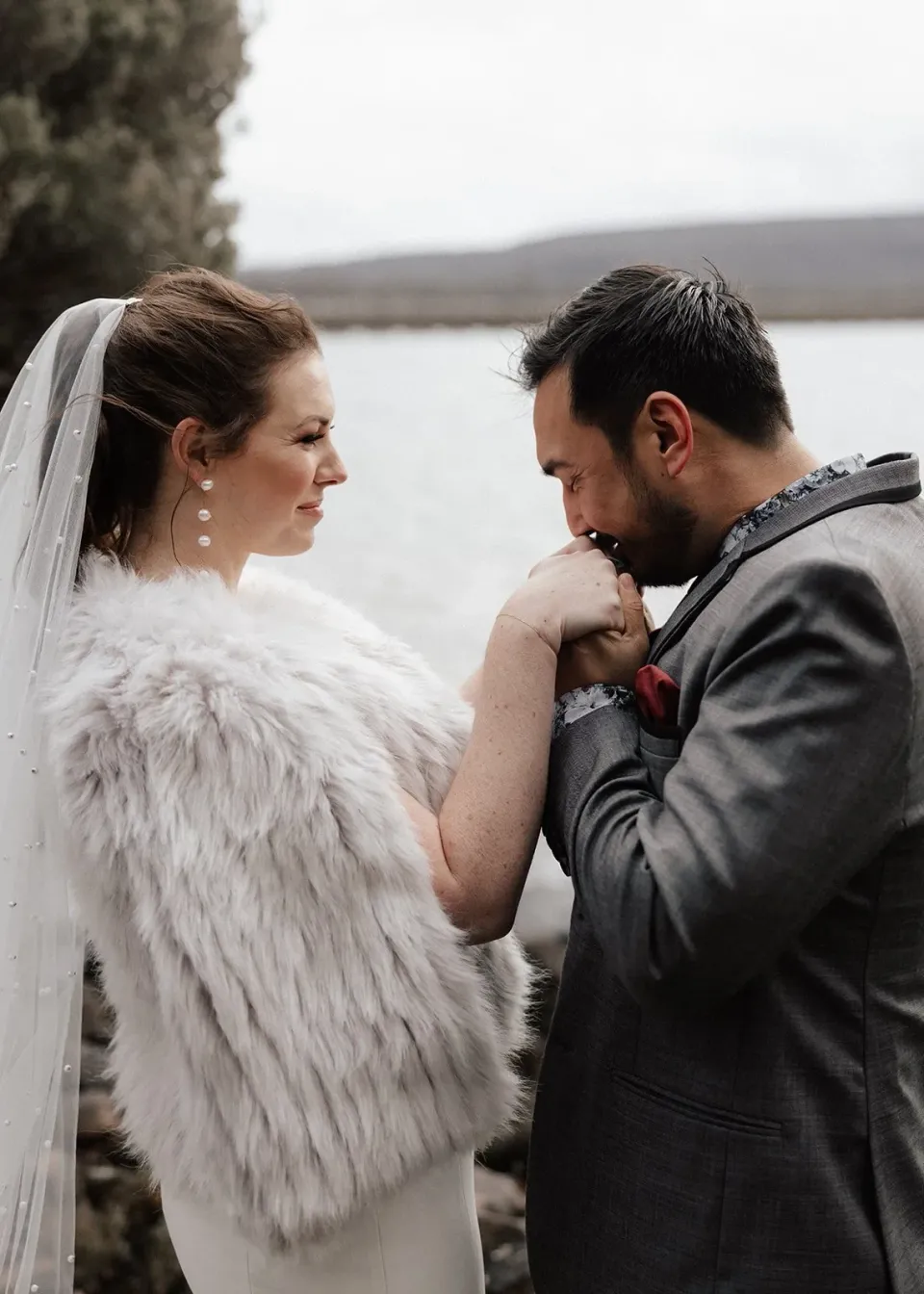 Jono kissing Sarah’s hand as she smiles beside the lake, at Pumphouse Point, Lake St Clair, Tasmania, for an Elopement Collective elopement. Photo by Bulb Creative.