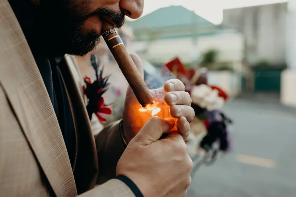 Close-up of Daniel lighting a cigar, with his beige suit and floral buttonhole visible, in Fortitude Valley, Brisbane, for an Elopement Collective elopement. Photo by Tessa Shannon.