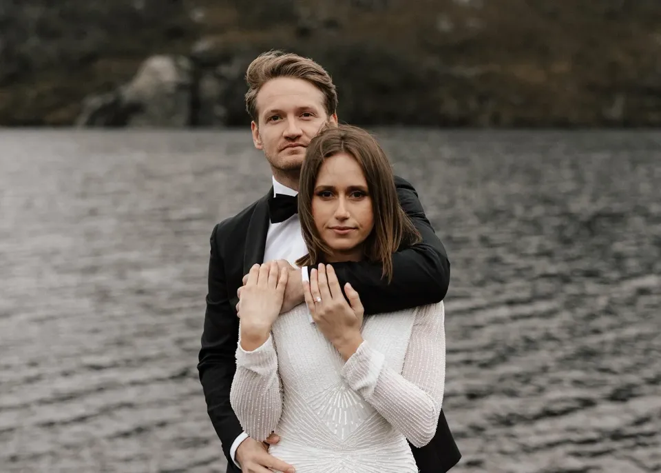 Josh embracing April from behind as she rests her hands on his arm beside the lake, at Dove Lake, Cradle Mountain, Tasmania, for an Elopement Collective elopement. Photo by Bulb Creative.