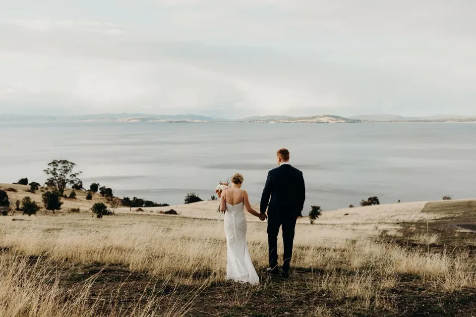 A couple walk through dry grass towards a wide bay beneath an overcast sky.