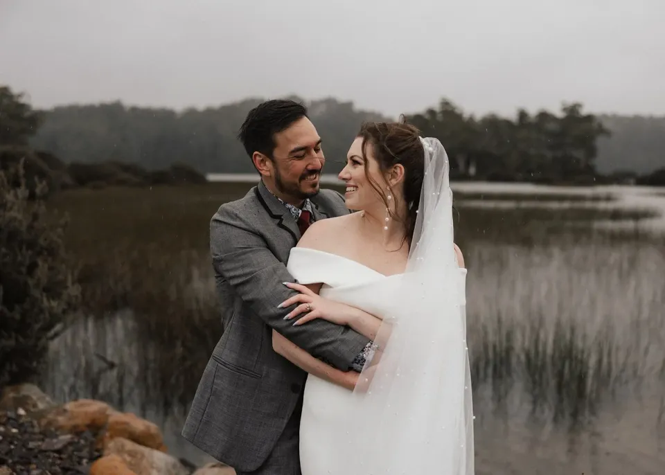 Jono embracing Sarah from behind beside lakeside reeds, with her off-the-shoulder white dress visible, at Pumphouse Point, Lake St Clair, Tasmania, for an Elopement Collective elopement. Photo by Bulb Creative.