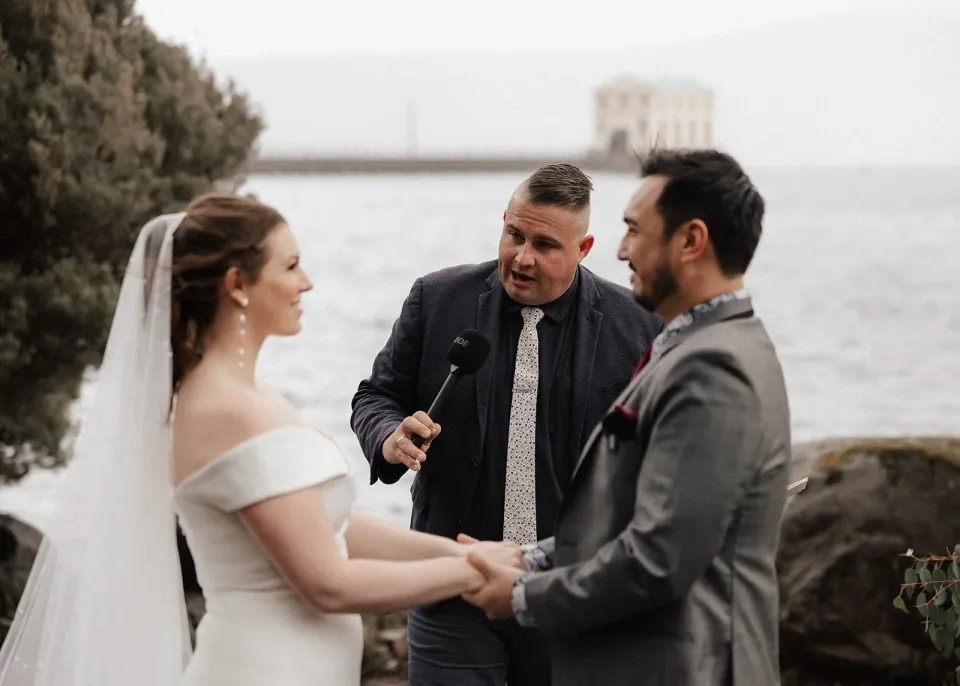 Sarah and Jono holding hands as celebrant Josh Withers speaks during their lakeside ceremony, at Pumphouse Point, Lake St Clair, Tasmania, for an Elopement Collective elopement. Photo by Bulb Creative.