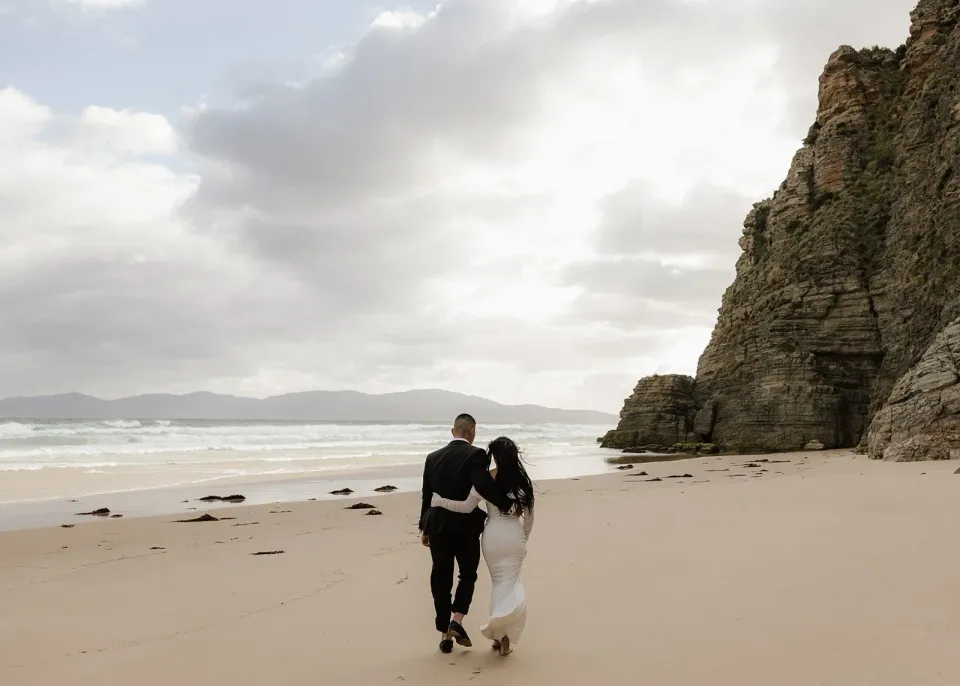 A couple walk along a sandy beach beneath towering rock cliffs and a cloudy sky.