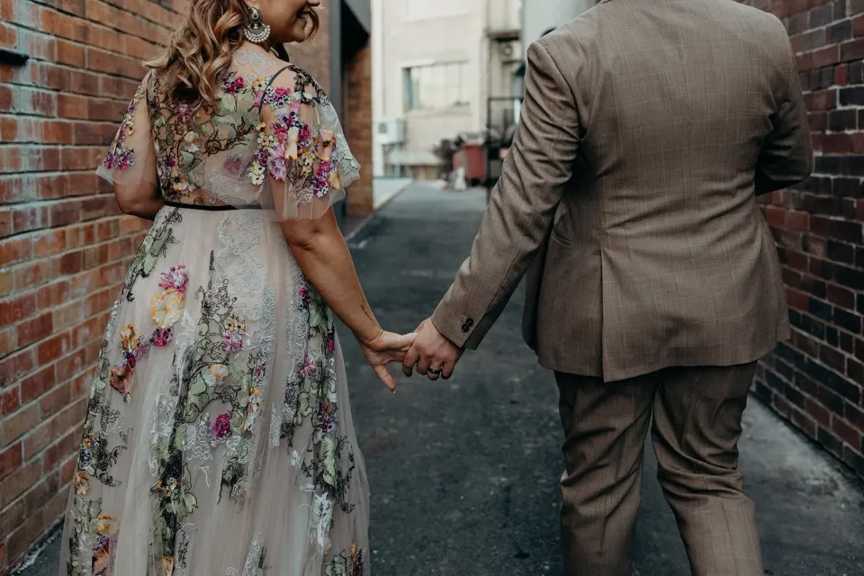 View from behind of Victoria and Daniel holding hands in a narrow brick-lined laneway, in Fortitude Valley, Brisbane, for an Elopement Collective elopement. Photo by Tessa Shannon.