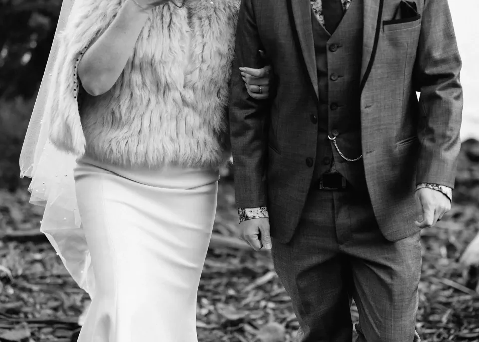 Black-and-white detail of Sarah’s pale textured wrap and white dress beside Jono’s grey three-piece suit as they walk arm in arm, at Pumphouse Point, Lake St Clair, Tasmania, for an Elopement Collective elopement. Photo by Bulb Creative.