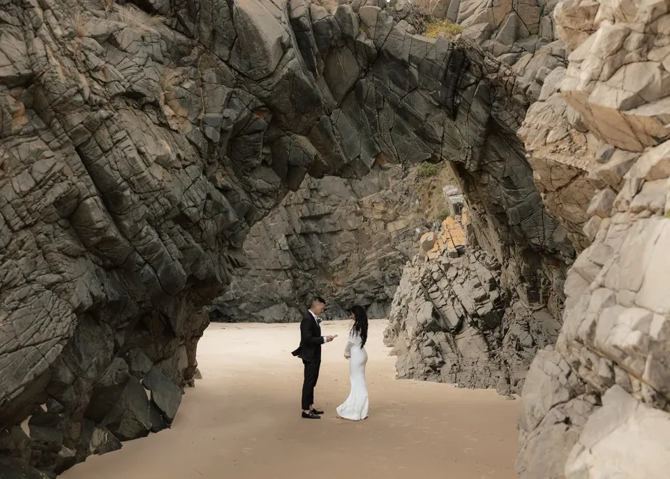 A couple stand on sand beneath a natural arch in towering coastal rock.
