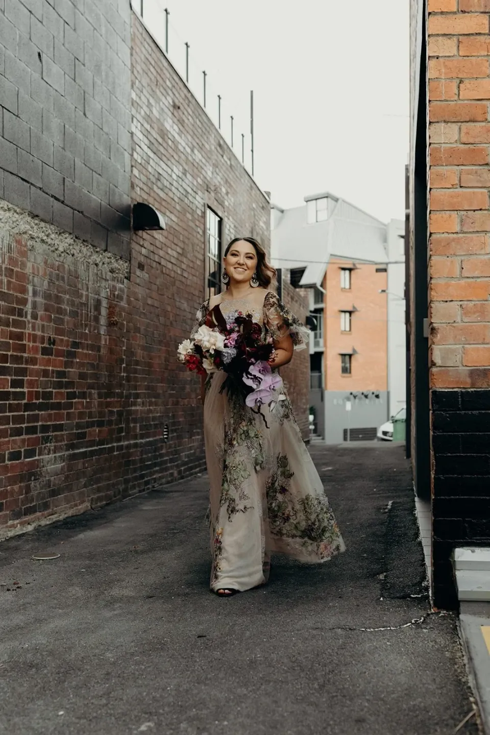 Full-length portrait of Victoria with her bouquet by Mrs Gibbons Flowers in a narrow brick-lined laneway, in Fortitude Valley, Brisbane, for an Elopement Collective elopement. Photo by Tessa Shannon.