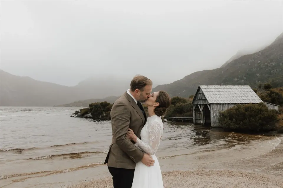 A couple kiss on a sandy lakeshore beside a timber boatshed and misty mountains.
