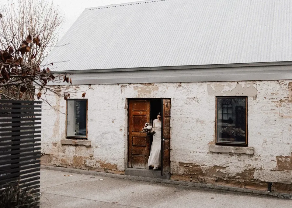 A bride stands in the doorway of a small, weathered stone cottage.