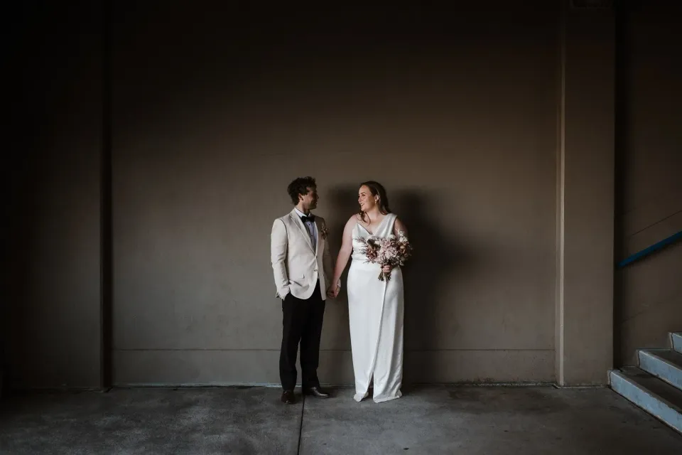 Alyce and Andrew looking at each other against a shadowed wall, her bouquet by The Lillipillian in hand, in The Rocks, Sydney, New South Wales, for an Elopement Collective elopement. Photo by Jason Corroto.