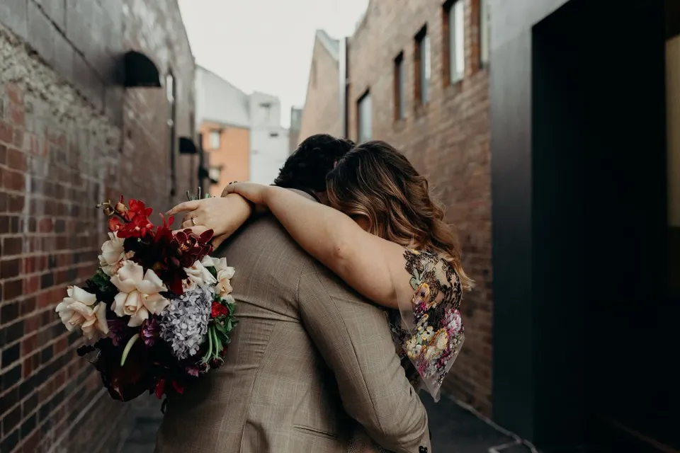 Victoria embracing Daniel with her arms around his shoulders and her bouquet by Mrs Gibbons Flowers behind his back, in Fortitude Valley, Brisbane, for an Elopement Collective elopement. Photo by Tessa Shannon.