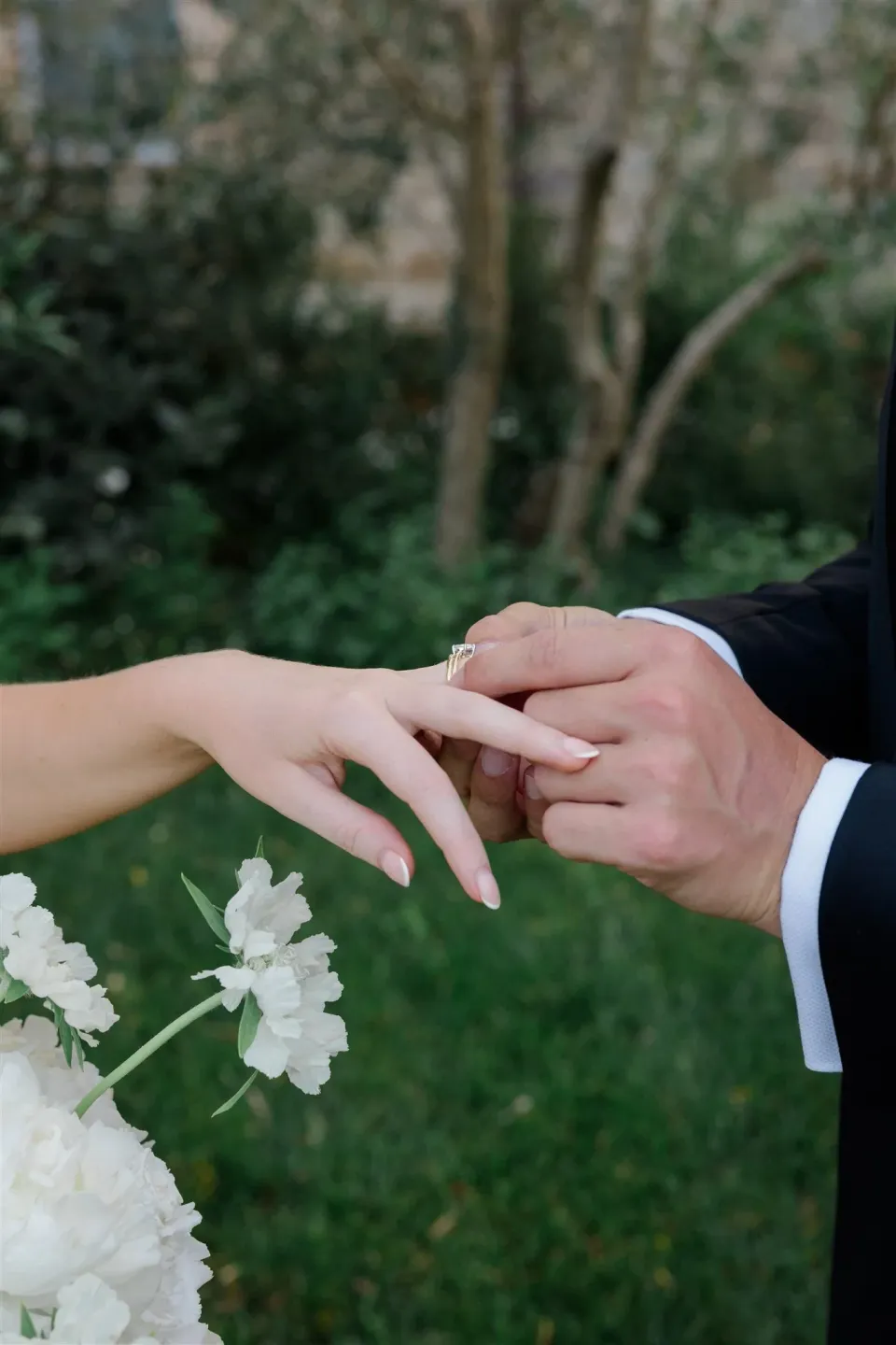 Close view of Jack placing a wedding ring on Shelby’s hand with white flowers by Le Francesche in the foreground, in Umbria, Italy, for an Elopement Collective elopement. Photo by James from House of Love.