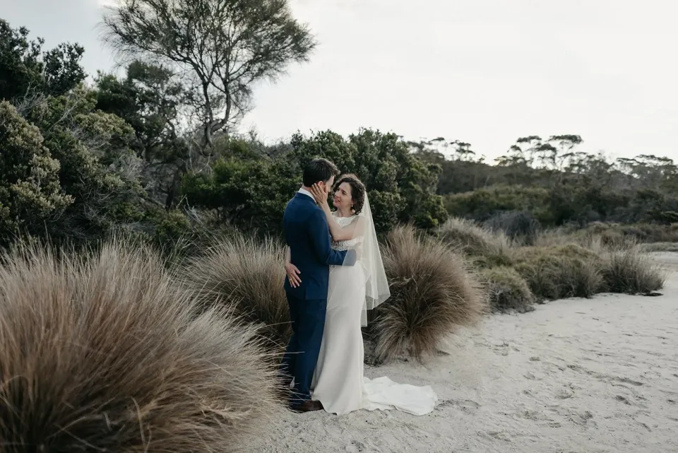 A couple embrace on white sand beside tussock grasses and coastal scrub.
