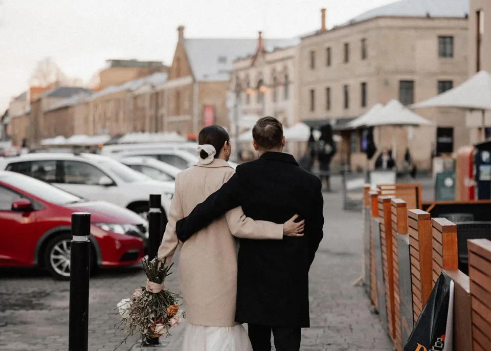 A couple walk arm in arm past sandstone buildings and outdoor tables on a Hobart street.