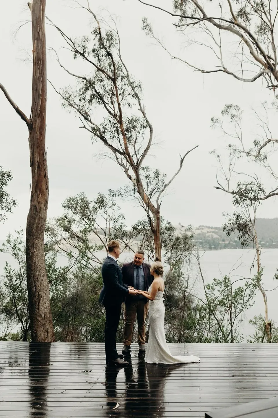 A couple stand together on a rain-darkened timber deck beside the water.