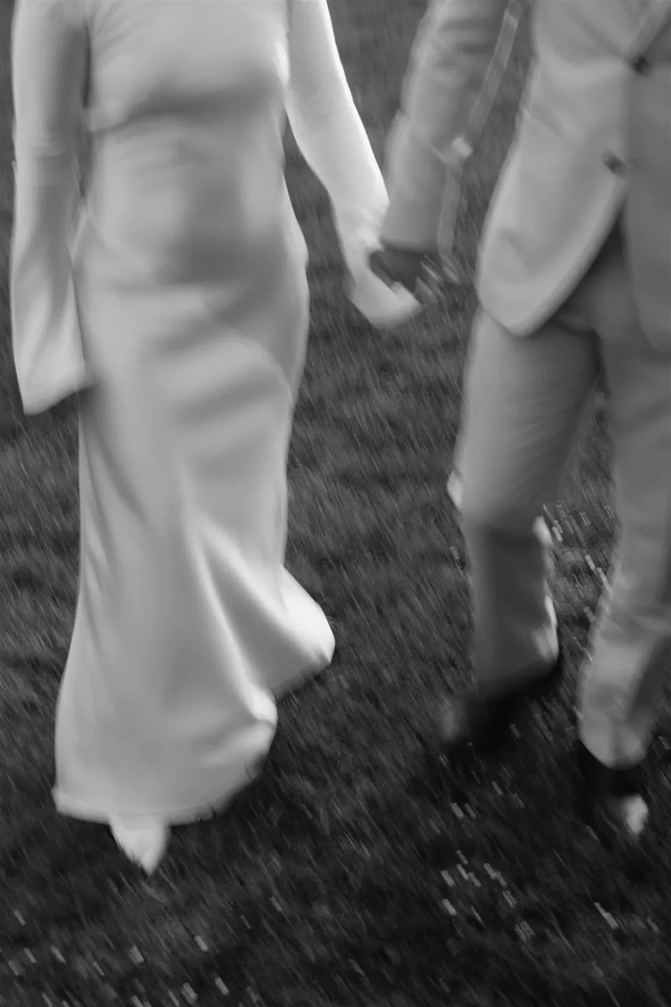 Black-and-white detail of Maddy’s dress and Cameron’s suit as they walk across the grass, at The Mayor’s Residence in Daylesford, Victoria, for an Elopement Collective elopement. Photo by Bulb Creative.