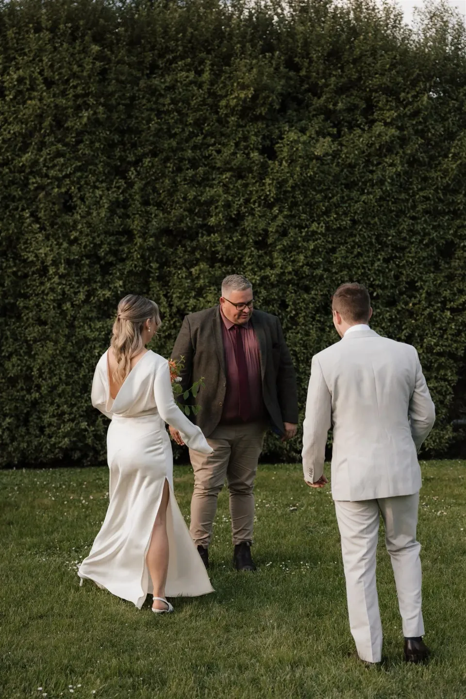 Maddy raising her hand to her face as she and Cameron stand with celebrant Josh Withers on the garden lawn, at The Mayor’s Residence in Daylesford, Victoria, for an Elopement Collective elopement. Photo by Bulb Creative.