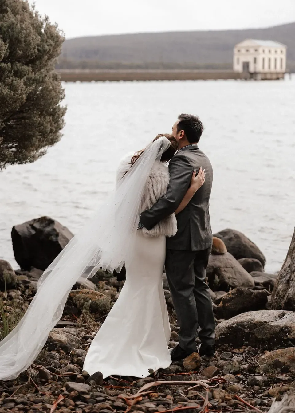 Sarah leaning back in Jono’s arms, her long veil lifting in the lakeside breeze, at Pumphouse Point, Lake St Clair, Tasmania, for an Elopement Collective elopement. Photo by Bulb Creative.