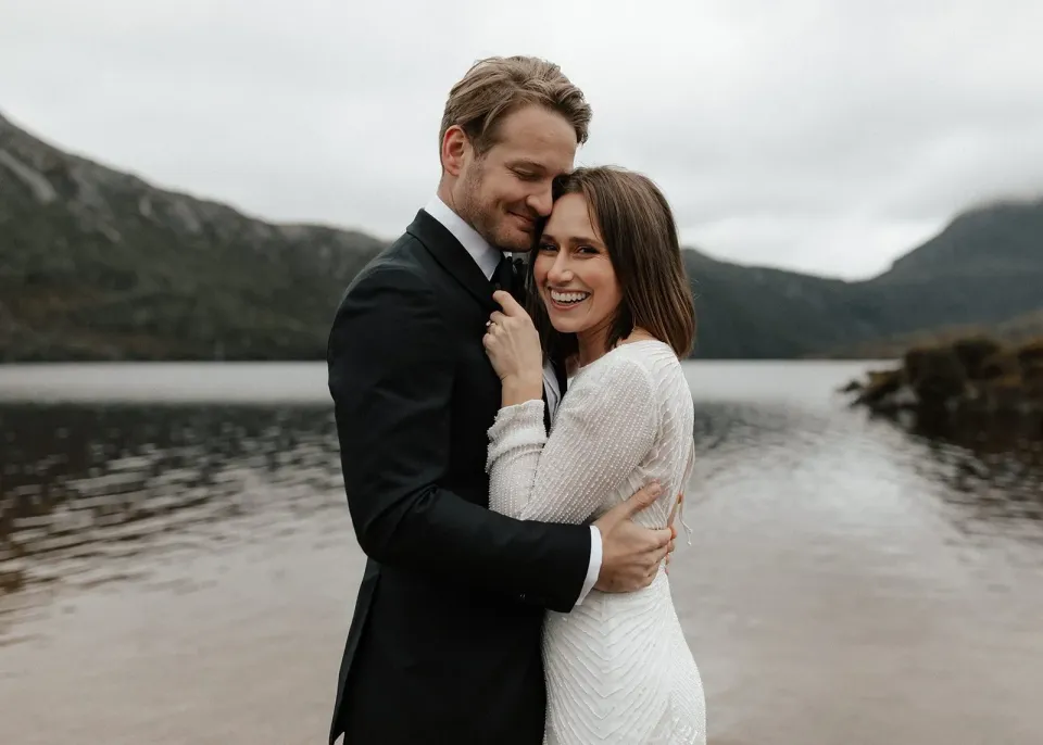 April laughing against Josh’s chest as he holds her close on the lakeshore, at Dove Lake, Cradle Mountain, Tasmania, for an Elopement Collective elopement. Photo by Bulb Creative.