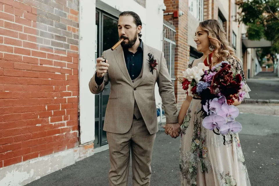 Daniel smoking a cigar as Victoria smiles beside him with her bouquet by Mrs Gibbons Flowers on a brick-lined street, in Fortitude Valley, Brisbane, for an Elopement Collective elopement. Photo by Tessa Shannon.