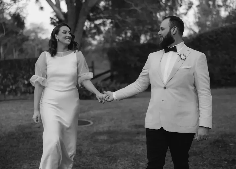 Black-and-white photograph of Tineeka and Chris smiling at each other while walking hand in hand, in Montville, Sunshine Coast Hinterland, Queensland, for an Elopement Collective elopement. Photo by Bulb Creative.