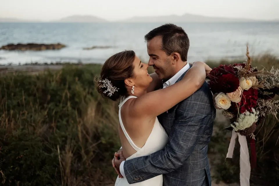 A bride embraces the groom with both arms around his neck beside the sea.