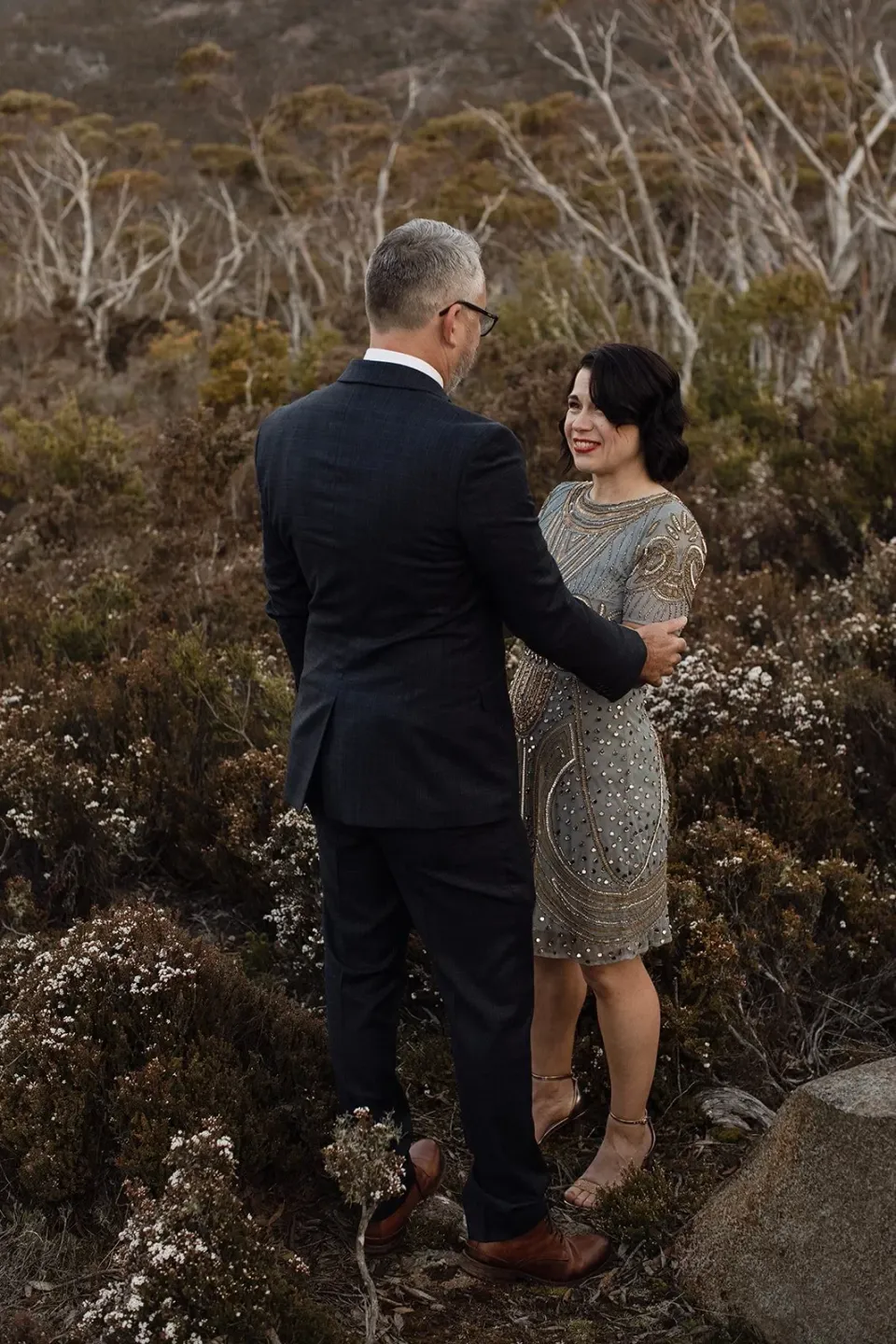 A couple face each other on a rocky path surrounded by mountain scrub.