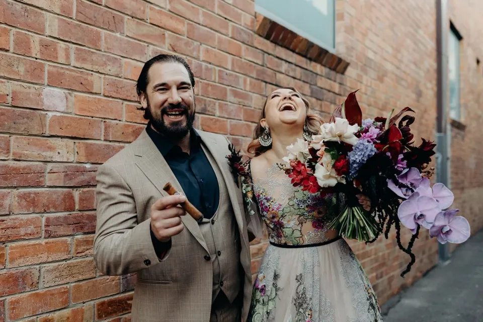 Victoria and Daniel laughing against a red brick wall, with her bouquet by Mrs Gibbons Flowers, in Fortitude Valley, Brisbane, for an Elopement Collective elopement. Photo by Tessa Shannon.