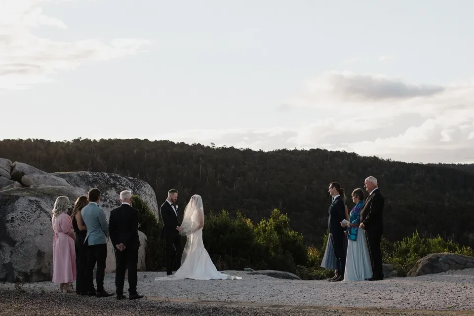 A small wedding party gathers around a couple for a ceremony on a rocky clearing.