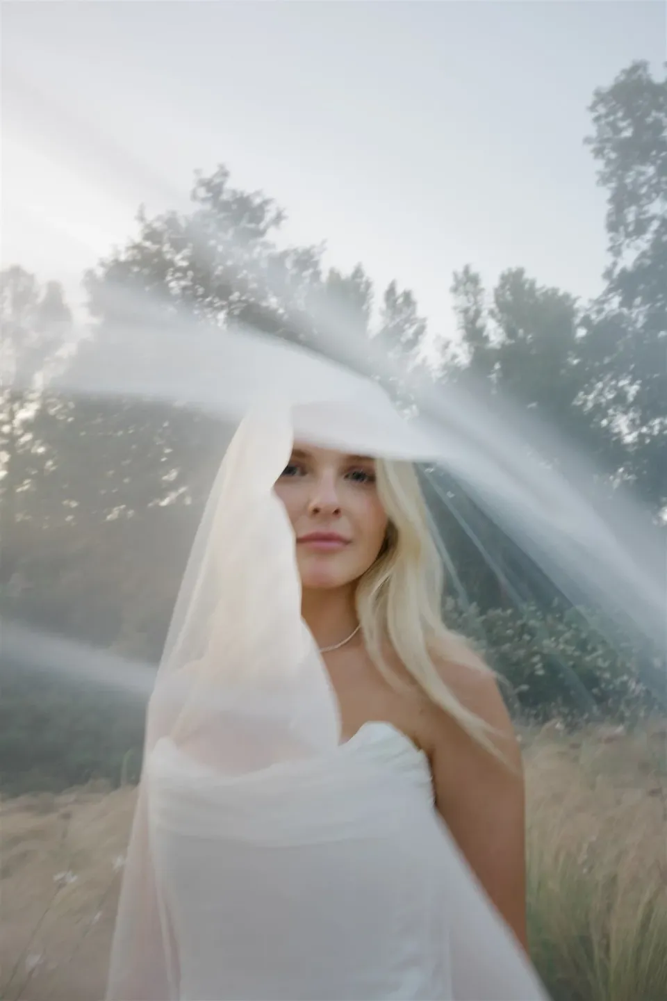 Shelby beneath her sheer veil, wearing her white Kate Willa dress among garden trees, in Umbria, Italy, for an Elopement Collective elopement. Photo by James from House of Love.