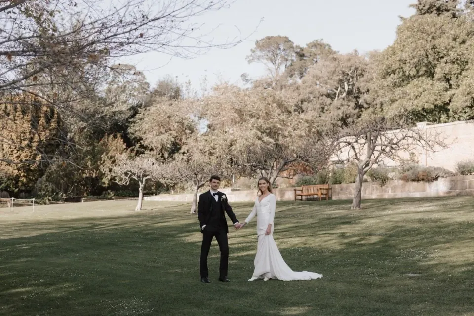 A couple hold hands across an open garden lawn bordered by trees.