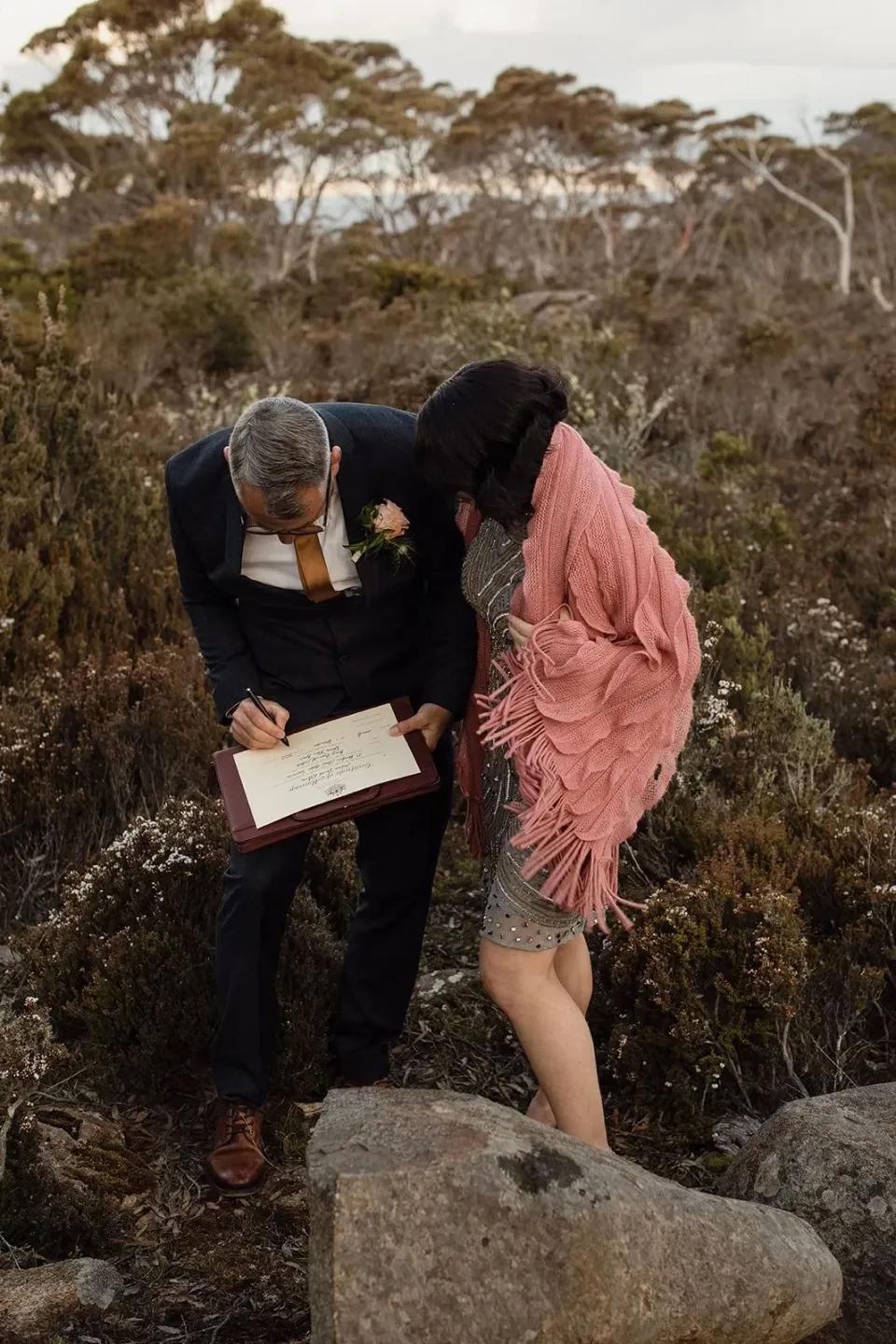 A couple lean over a document together among rocks and mountain scrub.