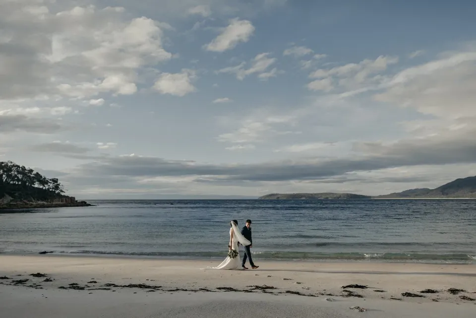 A couple walk along a quiet sandy beach beneath a wide, cloud-filled sky.