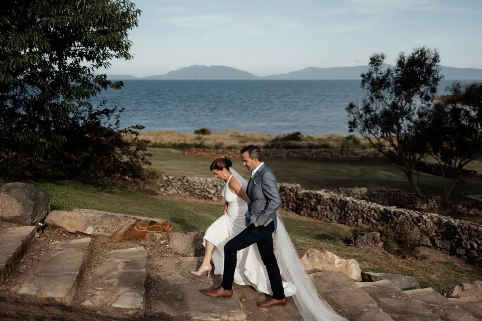 A couple climb worn stone steps beside a blue bay.
