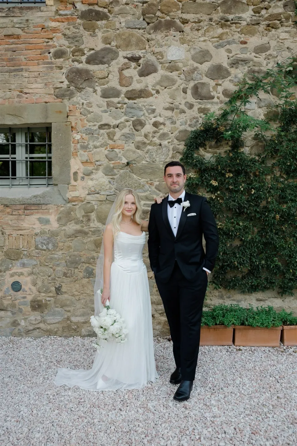 Shelby and Jack standing together on gravel in front of a vine-covered stone wall, in Umbria, Italy, for an Elopement Collective elopement. Photo by James from House of Love.