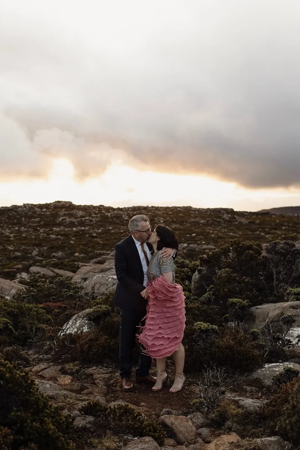 A couple stand close together among low alpine shrubs beneath a sunset sky.